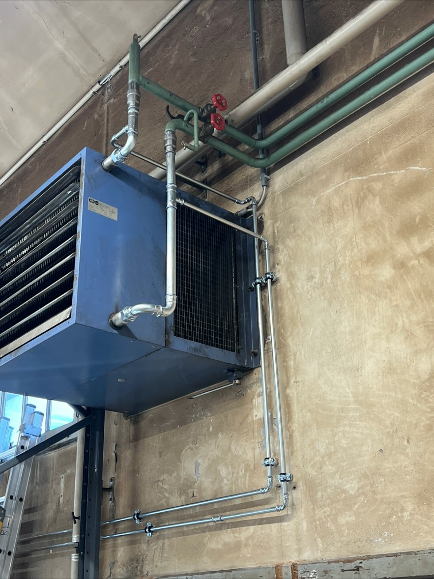 Hallenheizung – Warmlufterzeuger – Wandmontierter Warmlufterzeuger mit Rohrleitungsanschluss und Absperrventilen für eine Gewerbehalle.